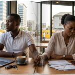 ADating couple sits silently at a café table, appearing emotionally distant as bills and financial documents lie between them, symbolizing money-related relationship stress.