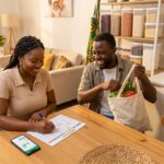 A happy Nigerian couple at a wooden dining table in a modern home. The woman with braided hair smiles while writing in a budgeting planner with a pen, as the man happily holds a canvas grocery bag filled with fresh vegetables. A smartphone showing financial charts sits on the table, illustrating smart family budgeting.