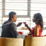 A stylishly dressed couple enjoys a romantic dinner, raising glasses of red wine during an elegant first date in a cozy restaurant with warm natural lighting.