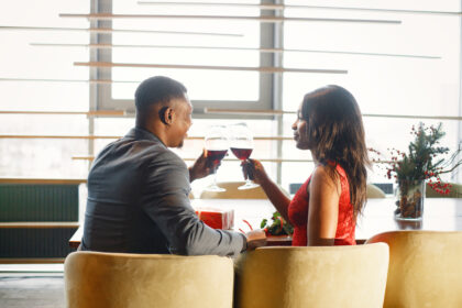 A stylishly dressed couple enjoys a romantic dinner, raising glasses of red wine during an elegant first date in a cozy restaurant with warm natural lighting.