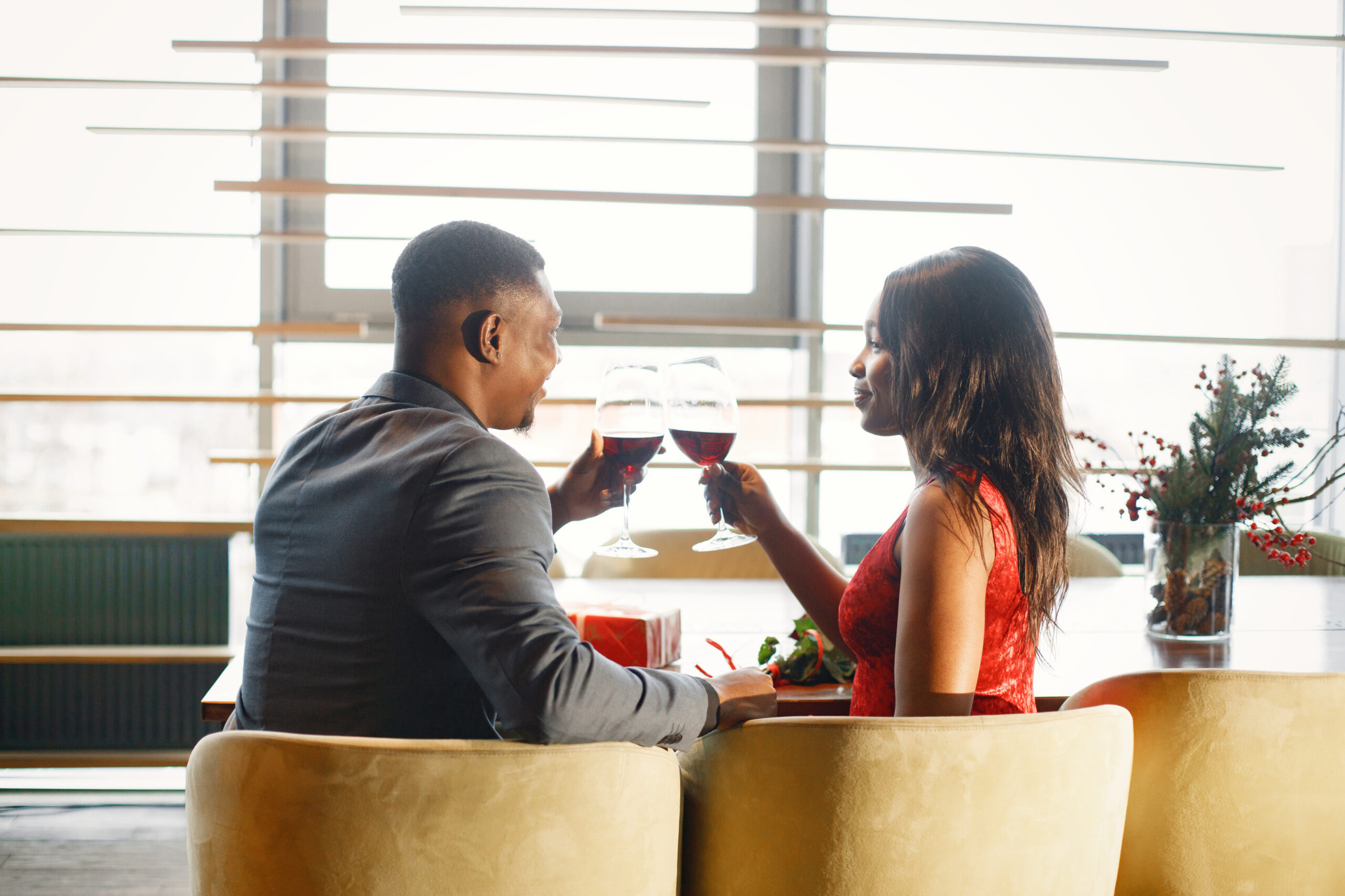 A stylishly dressed couple enjoys a romantic dinner, raising glasses of red wine during an elegant first date in a cozy restaurant with warm natural lighting.