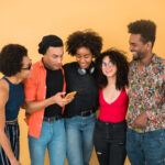 A joyful group of young Nigerian friends — representing Millennials and Gen Z — laughing and bonding together against a bright yellow wall as they happily view something on a smartphone.