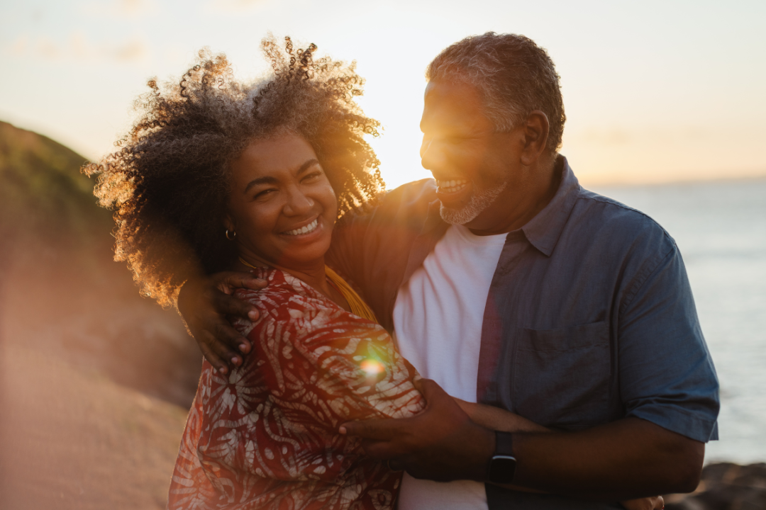 Smiling middle-aged couple embracing on a beach at sunset, sharing a warm and joyful moment that reflects trust, love, and emotional connection.