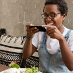 Young woman smiling while using a food delivery app on her smartphone, seated at a restaurant table with a healthy meal in front of her.