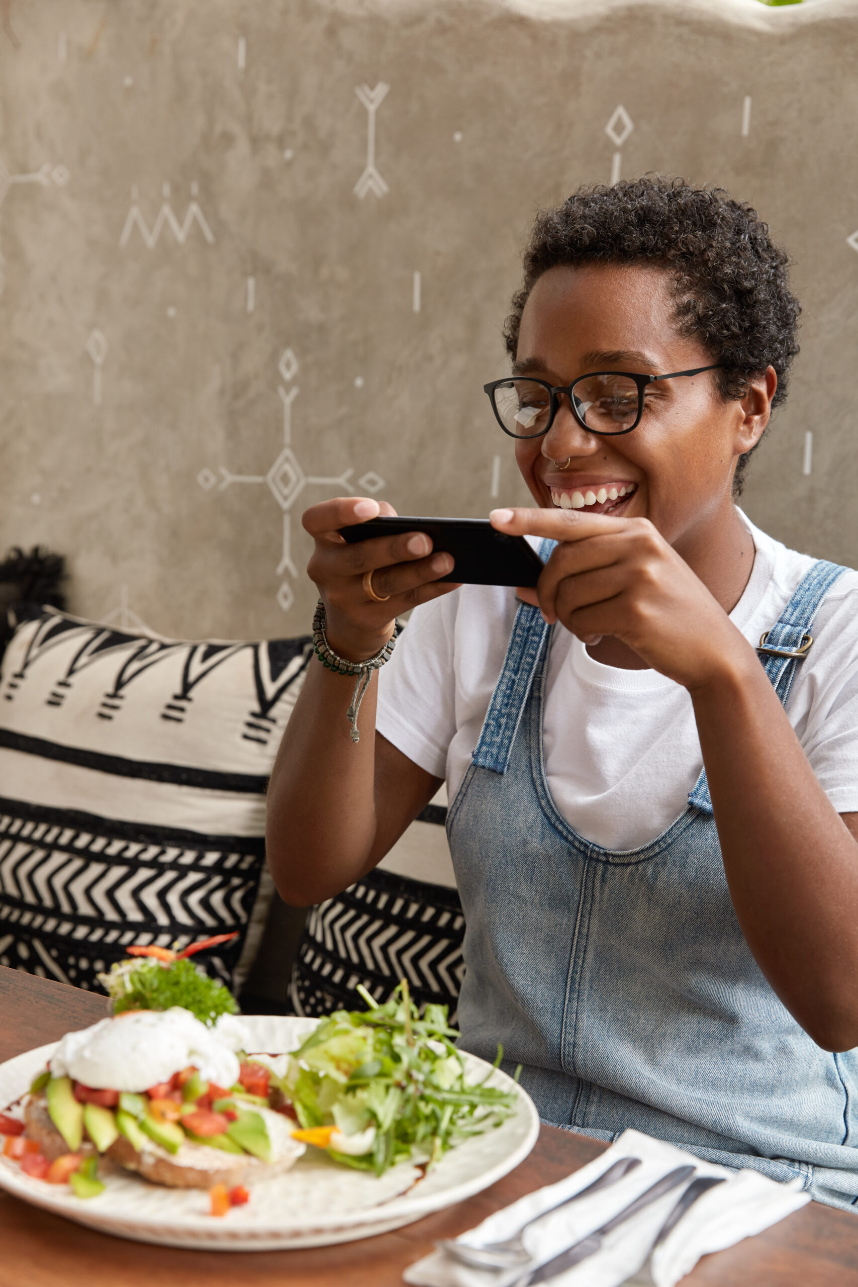 The Exciting Rise of Food Delivery Apps and their Impacts on Nigerian Eating Habits 12 Young woman smiling while using a food delivery app on her smartphone, seated at a restaurant table with a healthy meal in front of her.
