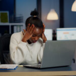 A stressed Black businesswoman holding her head in her hands while working late at night in a dimly lit office, with a laptop, documents, and a coffee mug on her desk.