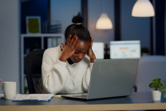 A stressed Black businesswoman holding her head in her hands while working late at night in a dimly lit office, with a laptop, documents, and a coffee mug on her desk.