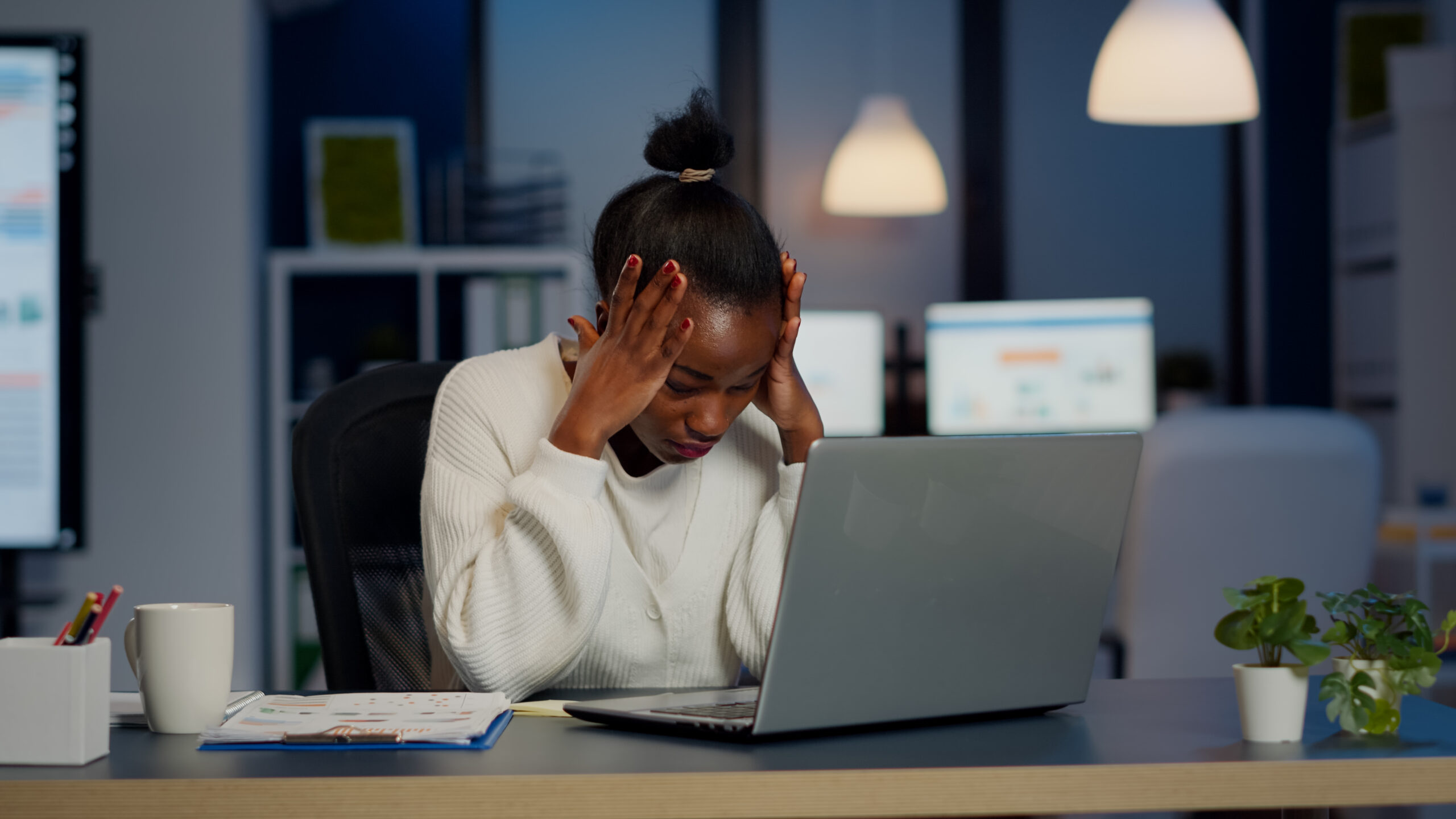 The Surprising Link Between City Life and Youth Burnout 12 A stressed Black businesswoman holding her head in her hands while working late at night in a dimly lit office, with a laptop, documents, and a coffee mug on her desk.