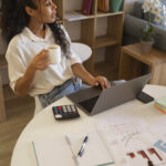 Young professional working from home on a laptop while reviewing financial charts and documents, holding a cup of coffee at a modern home office desk.