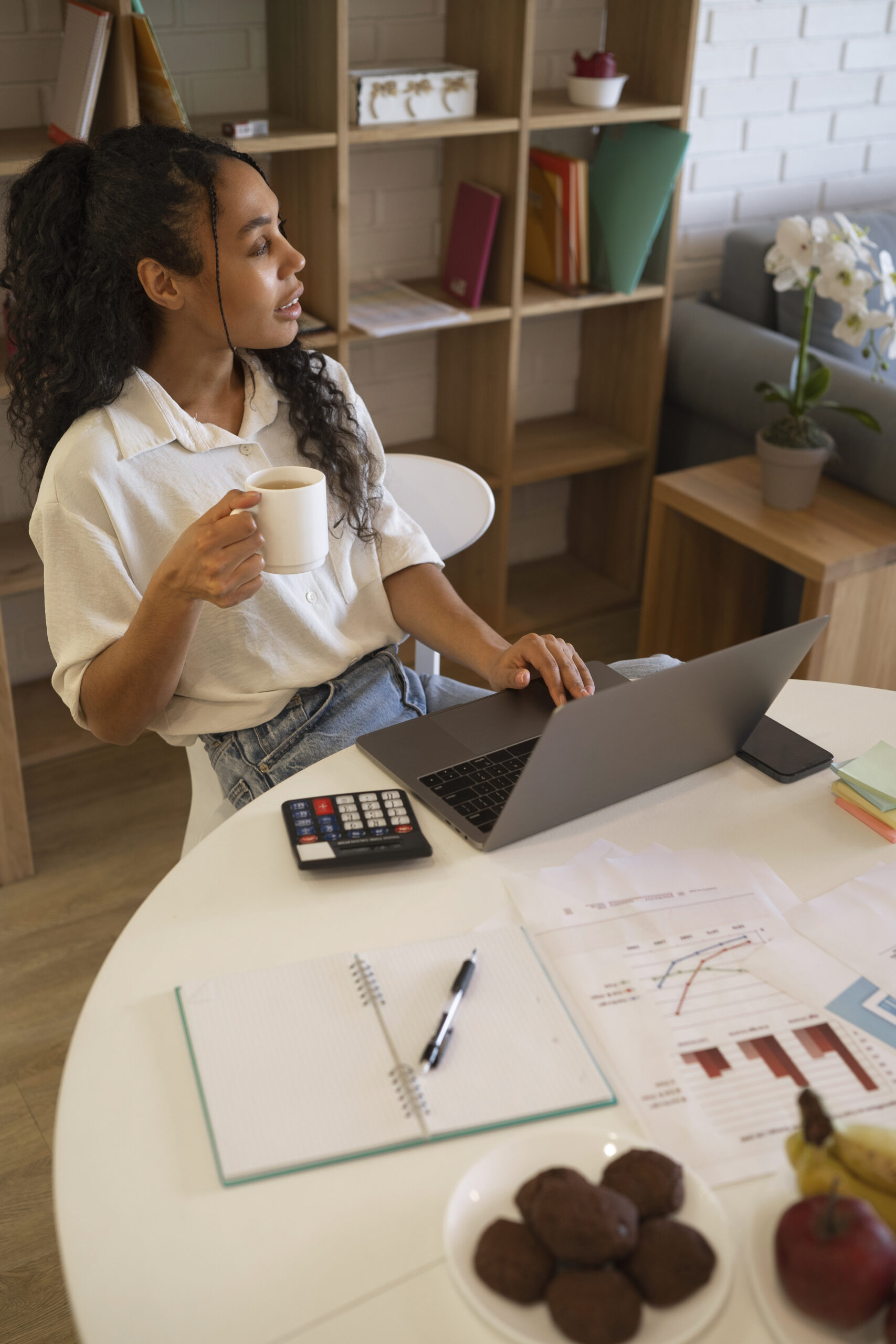 Young professional working from home on a laptop while reviewing financial charts and documents, holding a cup of coffee at a modern home office desk.
