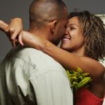 "Couple embracing closely indoors, woman in red dress holding flowers, smiling romantically"