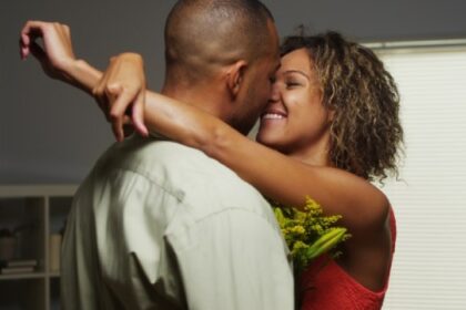 "Couple embracing closely indoors, woman in red dress holding flowers, smiling romantically"