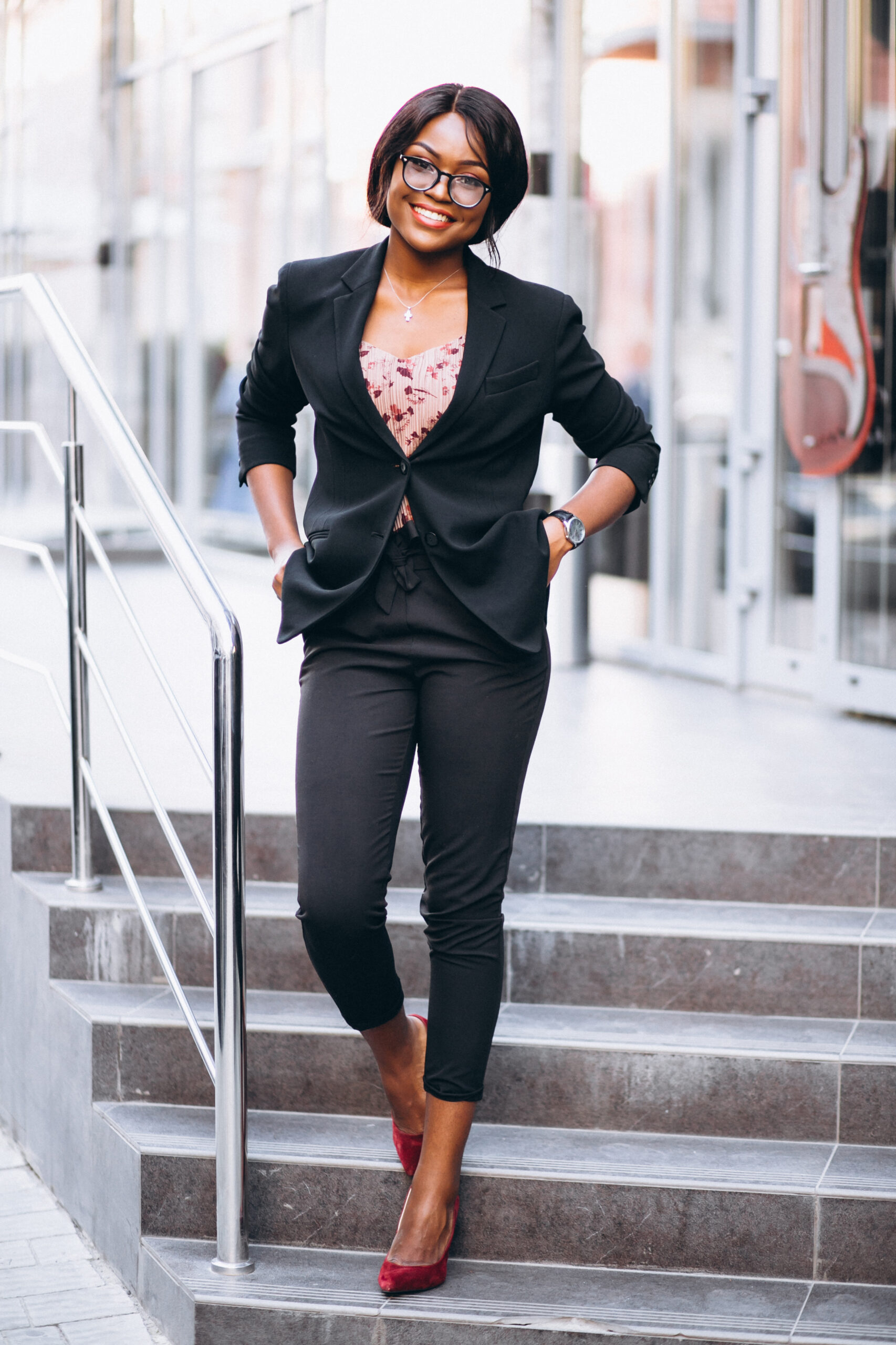 Young professional woman wearing a black blazer and tailored trousers with red heels, walking confidently down office steps in a stylish corporate outfit.