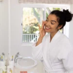Woman in a white bathrobe applying skincare in front of a bathroom mirror during her morning routine in a bright, modern home.